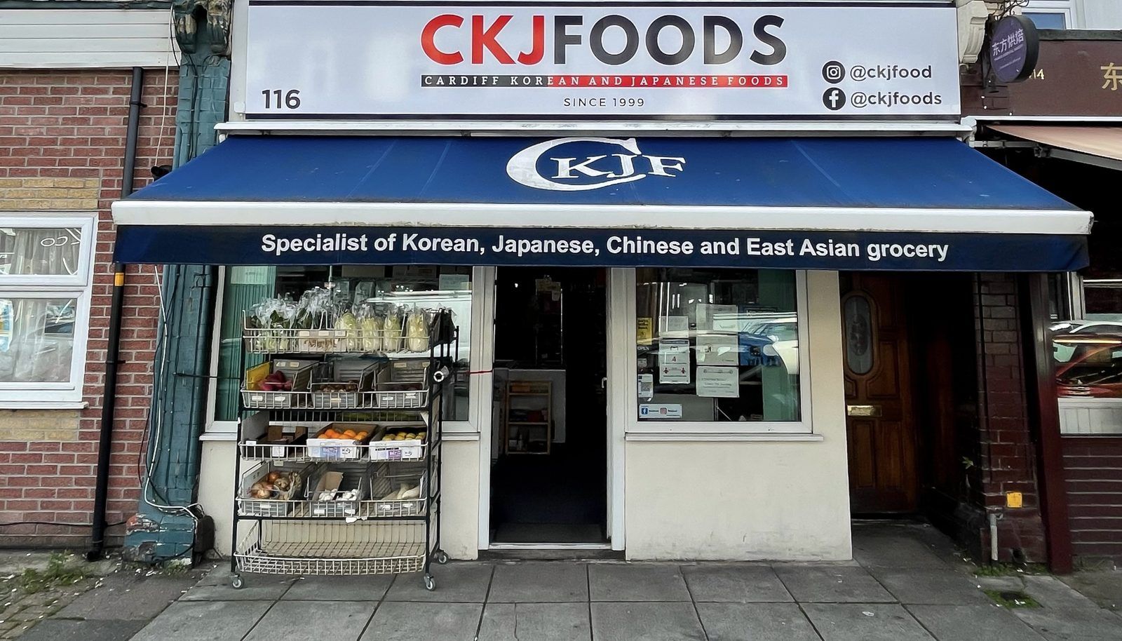 116번지의 CKJFoods 매장 외관. 빨강·남색의 CKJFOODS 간판, 파란색 CKJF 어닝, 태그라인 배너, 입구 앞에 진열된 신선 식재료가 보입니다